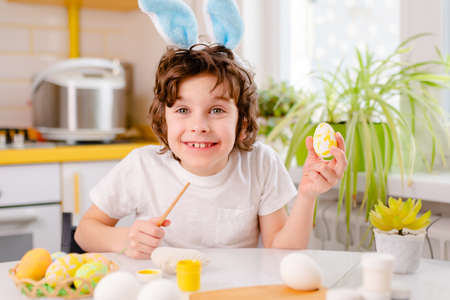 dyeing Easter eggs. curly boy dressed in white Tshirt and blue bunny ears paints Easter eggs.