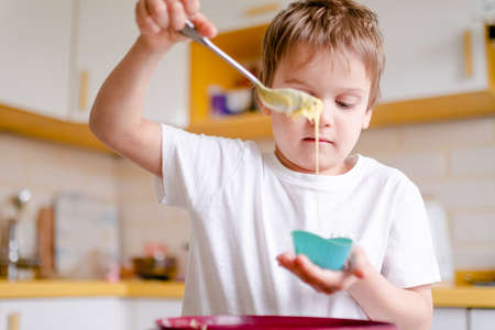 Little kid 5 years old cooks cupcakes in light kitchen at home. Little boy uses silicone baking...