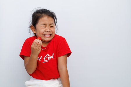 cute little asian girl with red t-shirt shows a sour expression after biting a sour-tasting strawberry. isolated on white background.の写真素材
