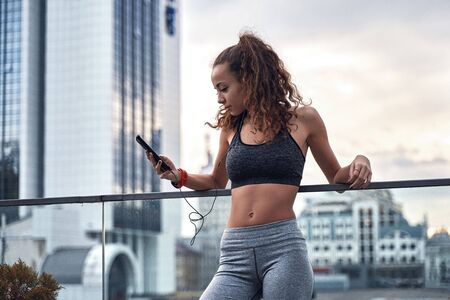 Young athletic woman in sport clothes standing on city background with phone in hands posting her...