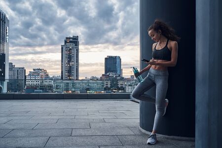 Young athletic woman in sport clothes standing on city background with phone in hands posting her...