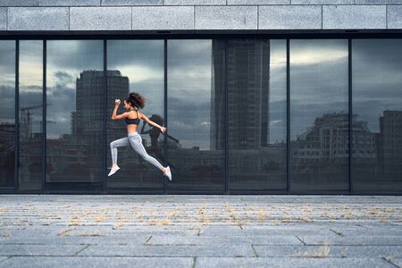 Young athletic woman jumping while making running training at urban city location with big mirror...