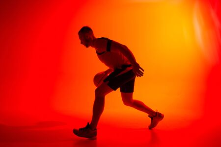 Young athletic man, basketball player dribbling with ball on red and orange backgroundの写真素材