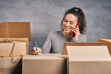 Woman writing addresses on the boxes, preparing parcels for send and receiving phone call. Working from homeの写真素材
