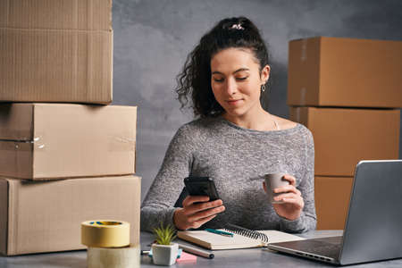 Business woman working from home with laptop and smartphone, preparing boxes with products for sendの写真素材