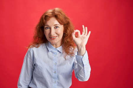 Smiling curly redhead woman showing Ok sign with her left hand on the red background