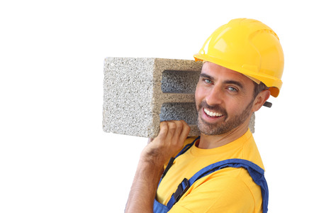 Confident handsome young builder or bricklayer wearing a safety helmet carrying a cement block on his shoulder and looking at the camera with a friendly smile, isolated on whiteの写真素材