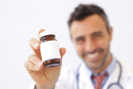 Smiling doctor holding up a bottle of tablets or pills with a blank white label for treatment of an illness or injury with focus to the bottleの写真素材