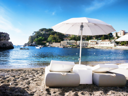 White elegant Sunbeds for romantic vacation at Mazzaro beach in Sicily Italy with hotels on the waterline and boats at the backgroundの写真素材