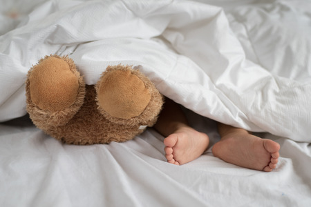 asian toddler feet beside teddy bear feet in white bed, sheet and pillow - toddler sleeping with teddy bearの写真素材