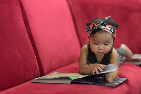 Asian baby toddler lying down on red sofa reading book. education conceptの写真素材