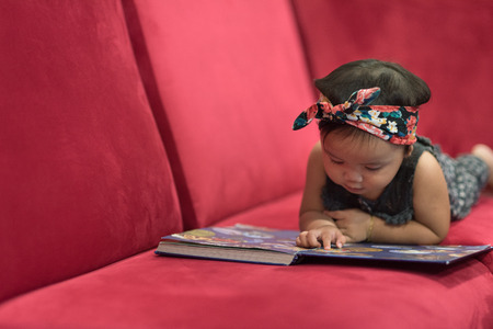 Asian baby toddler lying down on red sofa reading book. education concept