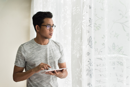 asian teen holding digital tablet while looking outside of his apartment. freelancer conceptの写真素材