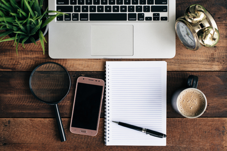 laptop, green plant, clock, magnifying glass, phone, coffee and blank notebook on a wooden table. work at home conceptの写真素材