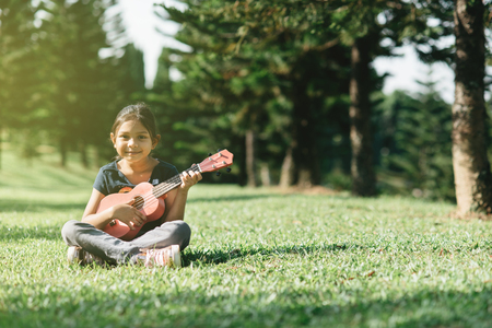 young and happy asian girl playing ukelele guitar in the park at sunny morning while looking at camera. hobbies and tranquility conceptの写真素材
