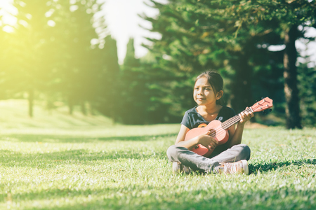 young and happy asian girl playing ukelele guitar in the park at sunny morning while looking to copy space. hobbies and tranquility conceptの写真素材
