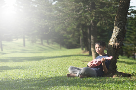 young asian boy having fun time playing ukelele at park in the sunny morningの写真素材