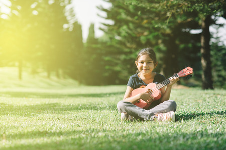 young and happy asian girl playing ukelele guitar in the park at sunny morning while looking at camera. hobbies and tranquility conceptの写真素材