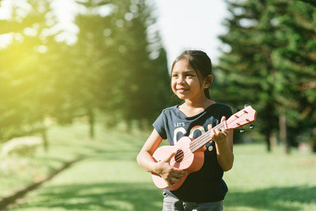 young and happy asian girl playing ukelele guitar in the park at sunny morning while looking to copy space. hobbies and tranquility conceptの写真素材