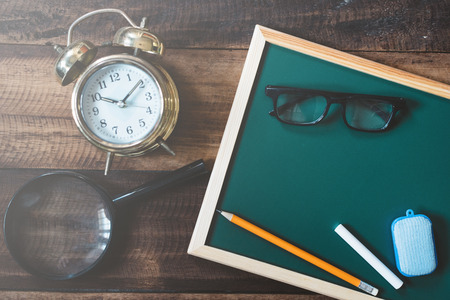 blank chalkboard with alarm clock,magnifying glass,eye glasses,pencil,white chalk and duster on wooden table, education,teacher,work and learning conceptの写真素材