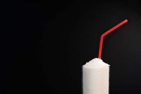 A glass full of white sugar with straw against black background. Concept of unhealthy eating and diabetisの写真素材