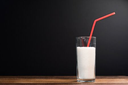 A glass full of white sugar with straw against black background. Concept of unhealthy eating and diabetisの写真素材