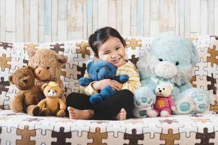 Cute little asian girl hugging her plush toy teddy bear on a sofa. Concept of childhood and happinessの写真素材