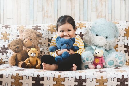 Cute little asian girl hugging her plush toy teddy bear on a sofa. Concept of childhood and happinessの写真素材
