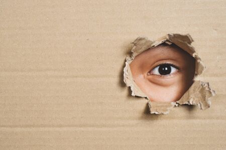 Boy eye peeking through a hole on a cardboard box with shocking gesture. Concept of mystery, secret and stalkerの写真素材