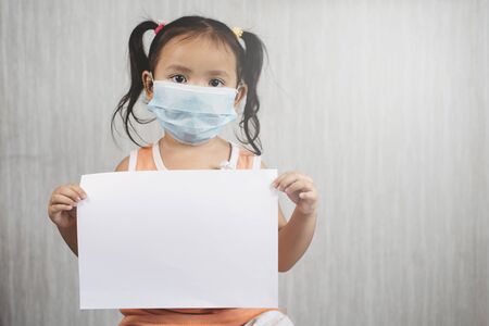 Little asian girl wearing surgical mask while holding blank white paper. Concept of child health care and virus desease outbreakの写真素材