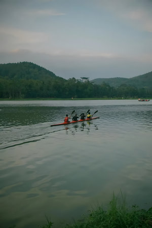 Group of people rowing a boat on a calm lake surrounded by lush green mountains under a cloudy sky