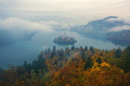 Amazing view of Lake Bled at foggy autumn morning with colorful forest on foreground and mountains covered by clouds on background, Sloveniaの写真素材