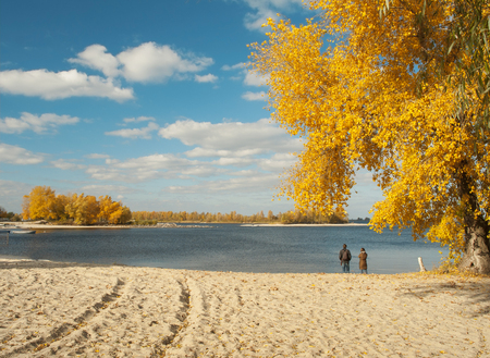 Autumn sunny landscape with autumn colorful trees, tracks in the sand and couple on river bankの写真素材