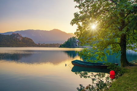 Magnificent sunrise over Lake Bled, Slovenia. Boat moored near lone tree. Sun shines through the tree crown. Bled Castle and Karavanke alps ridge in background. Travel destination sceneの写真素材