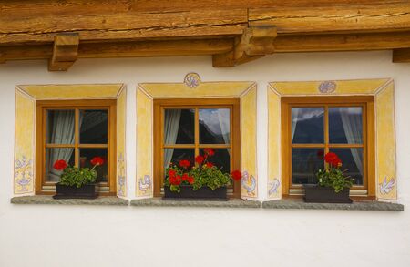 Three beautiful windows with red flowers and reflections of mountain in the glass in a small alpine country houseの写真素材