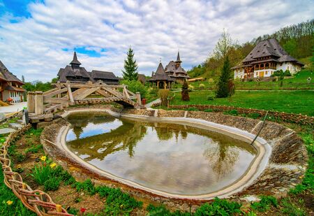 Decorative pond and wooden bridge in Barsana monastery, Romania. Traditional Maramures wooden architecture on background. Sunny spring day, Picturesque skyの写真素材