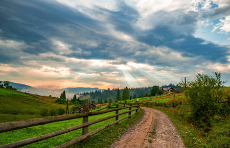 Breathtaking view of the sun makes its way through dramatic clouds and dirt road in Voronenko village in Carpathian National Park, Carpathian Mountains, Ukraine.の写真素材