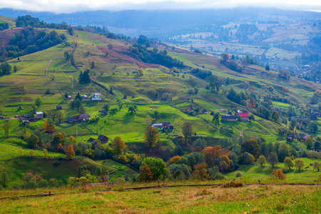 Scenic view of hills illuminated by autumnal sun in mountain village, Carpathian mountains, Lazeschyna, Ukraine. Yellowed trees on foreground and winding dirt road leading to top of the hillの写真素材