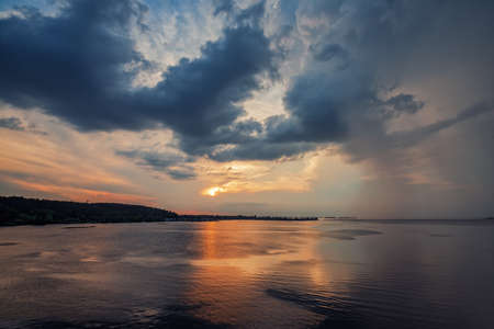 Dramatic sunset over Dnieper river near Cherkasy, Ukraine. Picturesque sky with dark clouds and sun path on wavy water. Scenic nature backgroundの写真素材
