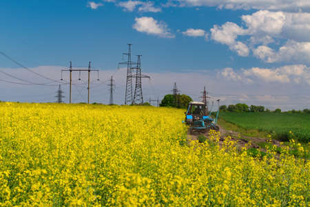 Tractor on blooming canola field. Power line support and high voltage electric line on background. agriculture conceptの写真素材