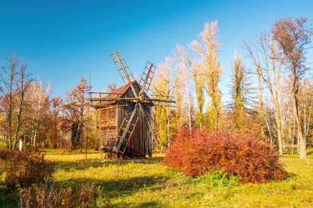 A picturesque autumn scene featuring a wooden windmill set in a rural landscape. The image captures...