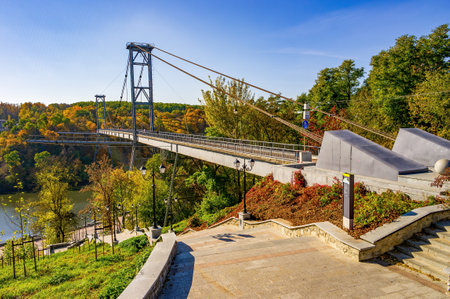 Picturesque view of a pedestrian suspension bridge crossing the Teteriv River in Zhytomyr, Ukraine. Steel cable blend structure harmoniously with autumn foliage of surrounding parkの写真素材