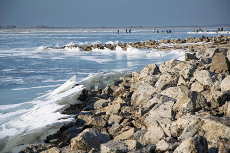 Thin ice sheets are accumulating on a rocky shore with people walking on a frozen lake in the background on a sunny winter dayの写真素材