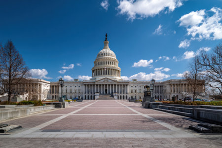 A grand building with a central dome is presented under a vibrant blue sky dotted with clouds. The architecture features classical lines, columns, and a symmetrical composition. The image showcases a daytime scene, suggesting a governmental or institutional environment, and is suitable for diverse commercial and editorial applications.の素材