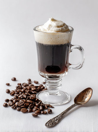 A transparent glass cup contains Irish coffee topped with whipped cream. Beside it, scattered coffee beans and a silver spoon lie on a clean white surface. The beverage is in a studio-style, well-lit environment suitable for drink-related publications.の素材