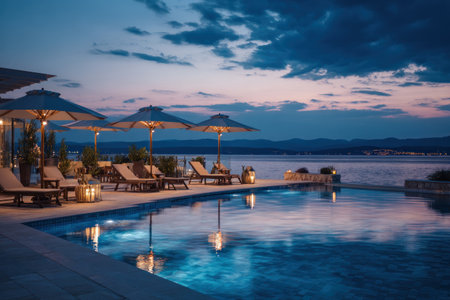 An outdoor scene depicts a pool, lounge chairs, and umbrellas overlooking a body of water at dusk. The composition showcases reflections and a dark blue color palette. Soft lighting accents the scene, hinting at a relaxing environment. This image can be used in various commercial projects, including travel and hospitality content.の素材