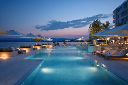 A serene scene showcases a long, rectangular swimming pool reflecting the twilight sky. Soft lighting illuminates the pool area, with lounge chairs and umbrellas adding elements of comfort. The architectural design of the surroundings suggests a luxurious resort setting, possibly suitable for hospitality and lifestyle content.の素材