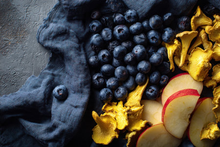 An overhead composition showcases fresh blueberries, sliced apples, and golden chanterelle mushrooms arranged on a textured blue cloth. The arrangement features a dark aesthetic with the vivid colors of the ingredients. This image could be suitable for commercial purposes such as food blogs, cookbooks, or product promotions.の素材