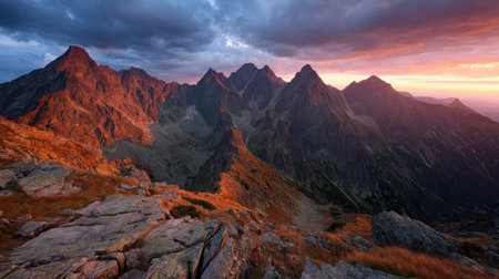 A stunning mountain range is illuminated by the warm hues of sunset, creating a dramatic contrast with the cloudy sky. The composition highlights rugged peaks and valleys. The scene evokes a sense of vastness and natural beauty, suitable for various commercial applications.の素材