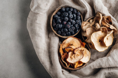 An overhead shot presents three small wooden bowls filled with dried apple slices and fresh blueberries. The bowls rest upon a crumpled, textured cloth in neutral tones. The lighting is soft and natural, creating a clean, minimalist aesthetic suitable for various food-related editorials.の素材
